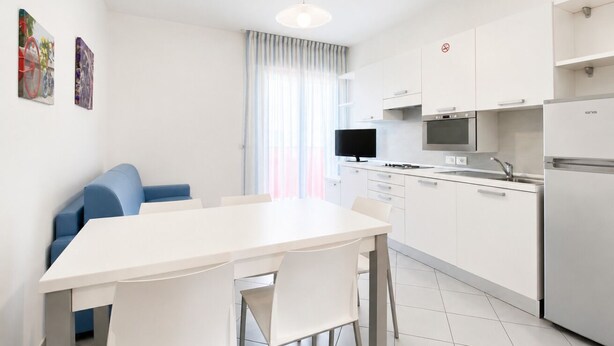 A modern kitchen with white cabinets, a dining table, and chairs. There is a television on the wall and a refrigerator on the right.
