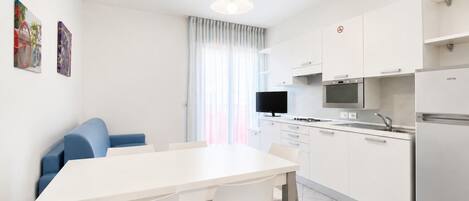A modern kitchen with white cabinets, a dining table, and chairs. There is a television on the wall and a refrigerator on the right.