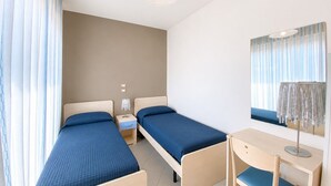 A hotel room with two single beds, a desk with a chair, a mirror, and a window with curtains.