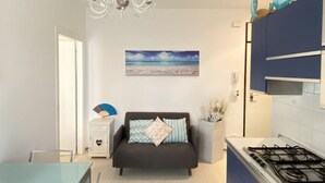 A modern kitchen with a gas stove, a dining area with a table and chairs, a sofa with cushions, and a decorative fan. - Appartamento Tarvisium - Carraro Immobiliare Jesolo (Lido di Jesolo)