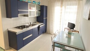A modern kitchen with dark blue cabinets, a gas stove, and a dining area with a glass table and chairs. - Appartamento Tarvisium - Carraro Immobiliare Jesolo (Lido di Jesolo)