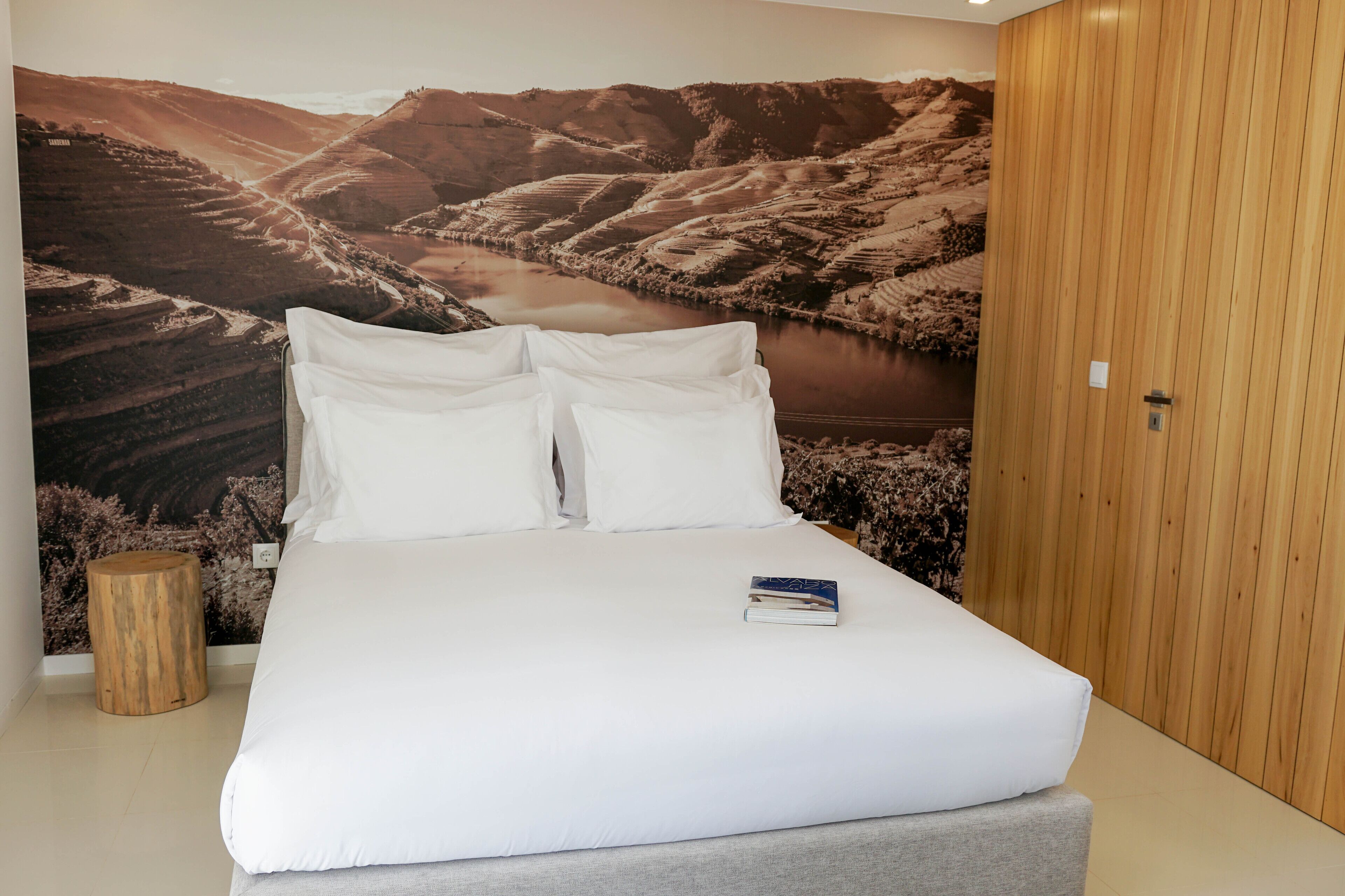 Una cama bien hecha con ropa blanca y almohadas, un libro sobre la cama y un amplio mural paisajístico en la pared de fondo.
