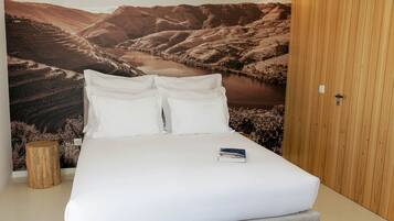 Una cama bien hecha con ropa blanca y almohadas, un libro sobre la cama y un amplio mural paisajístico en la pared de fondo.