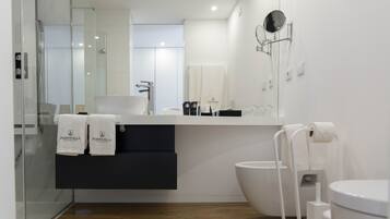 A modern bathroom with a glass shower enclosure, a sink with a black cabinet below, a toilet with a bidet attachment, and a mirror with a round frame.