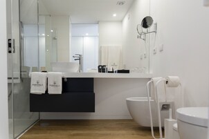 A modern bathroom with a glass shower enclosure, a sink with a black cabinet below, a toilet with a bidet attachment, and a mirror with a round frame.