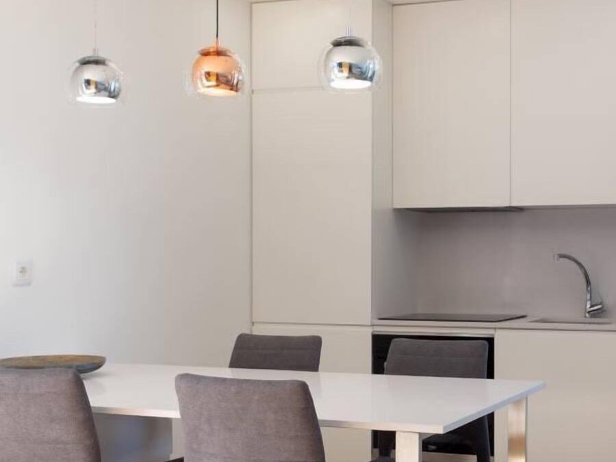 Una cocina moderna con armarios blancos, un lavamanos de acero inoxidable y una zona de comedor con una mesa blanca y sillas grises.