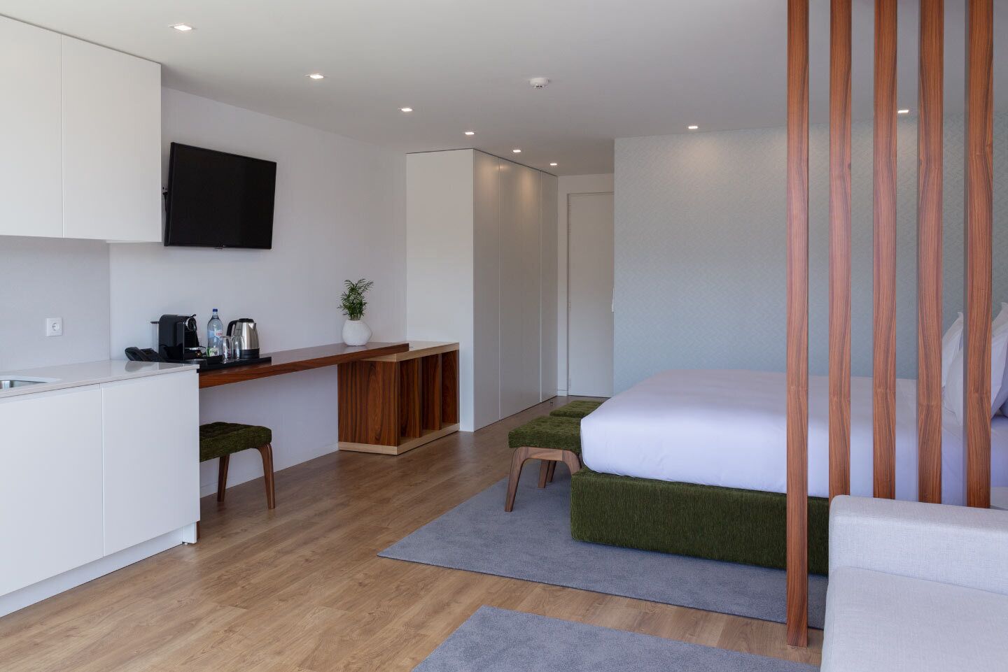 A modern, minimalist hotel room with a wooden desk, a bed with green upholstered headboard, and a flat-screen TV mounted on the wall.