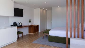 A modern, minimalist hotel room with a wooden desk, a bed with green upholstered headboard, and a flat-screen TV mounted on the wall.