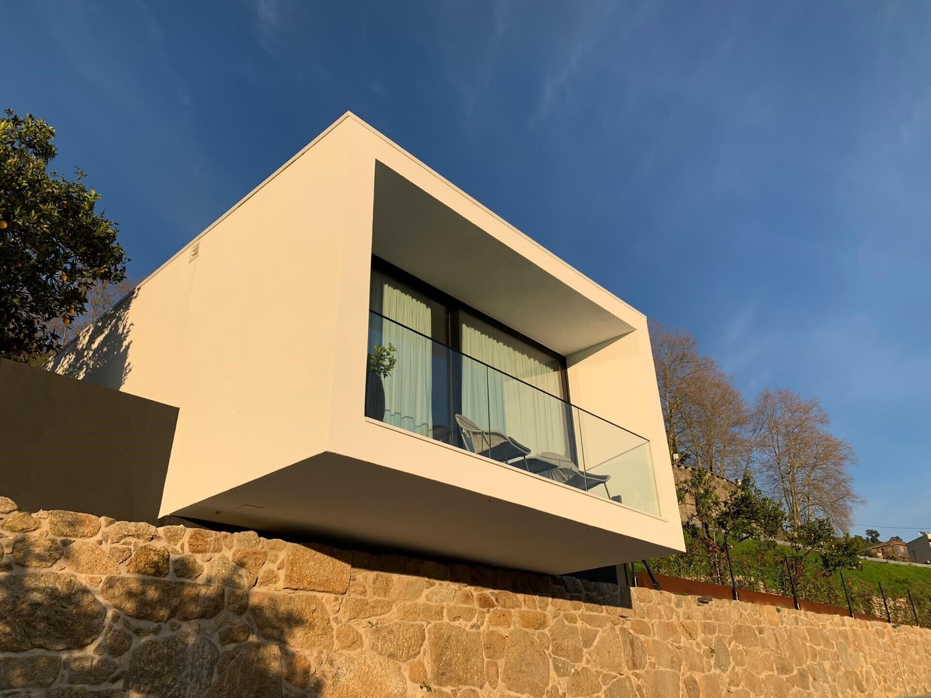 Una casa moderna con techo plano, un balcón con barandilla de vidrio y base de pared de piedra.