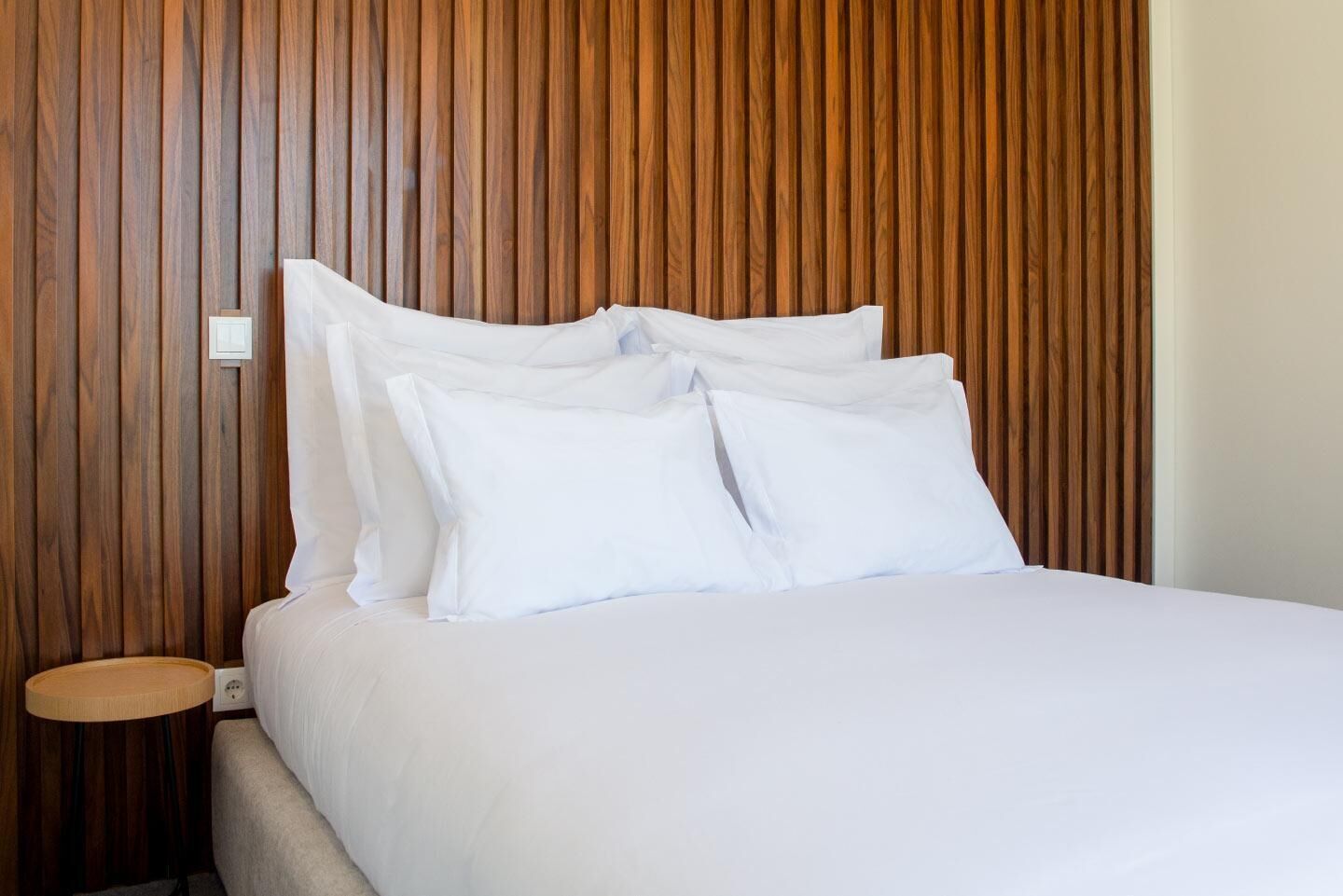 Een opgemaakt bed met witte lakens en kussens, een houten hoofdbord en een klein nachtkastje met een lamp.