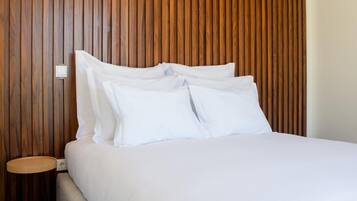 Een opgemaakt bed met witte lakens en kussens, een houten hoofdbord en een klein nachtkastje met een lamp.