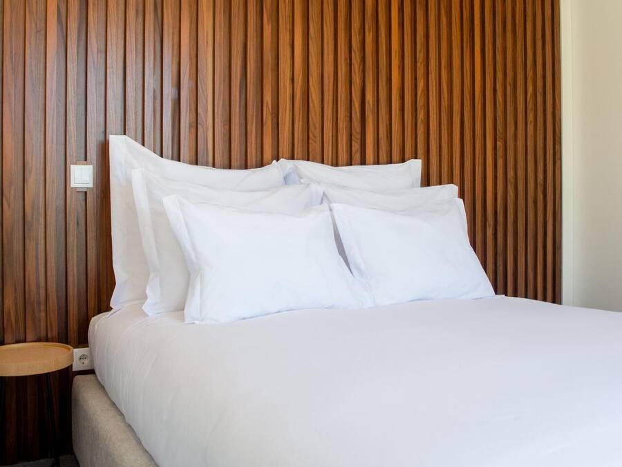 Een opgemaakt bed met witte lakens en kussens, een houten hoofdbord en een klein nachtkastje met een lamp.