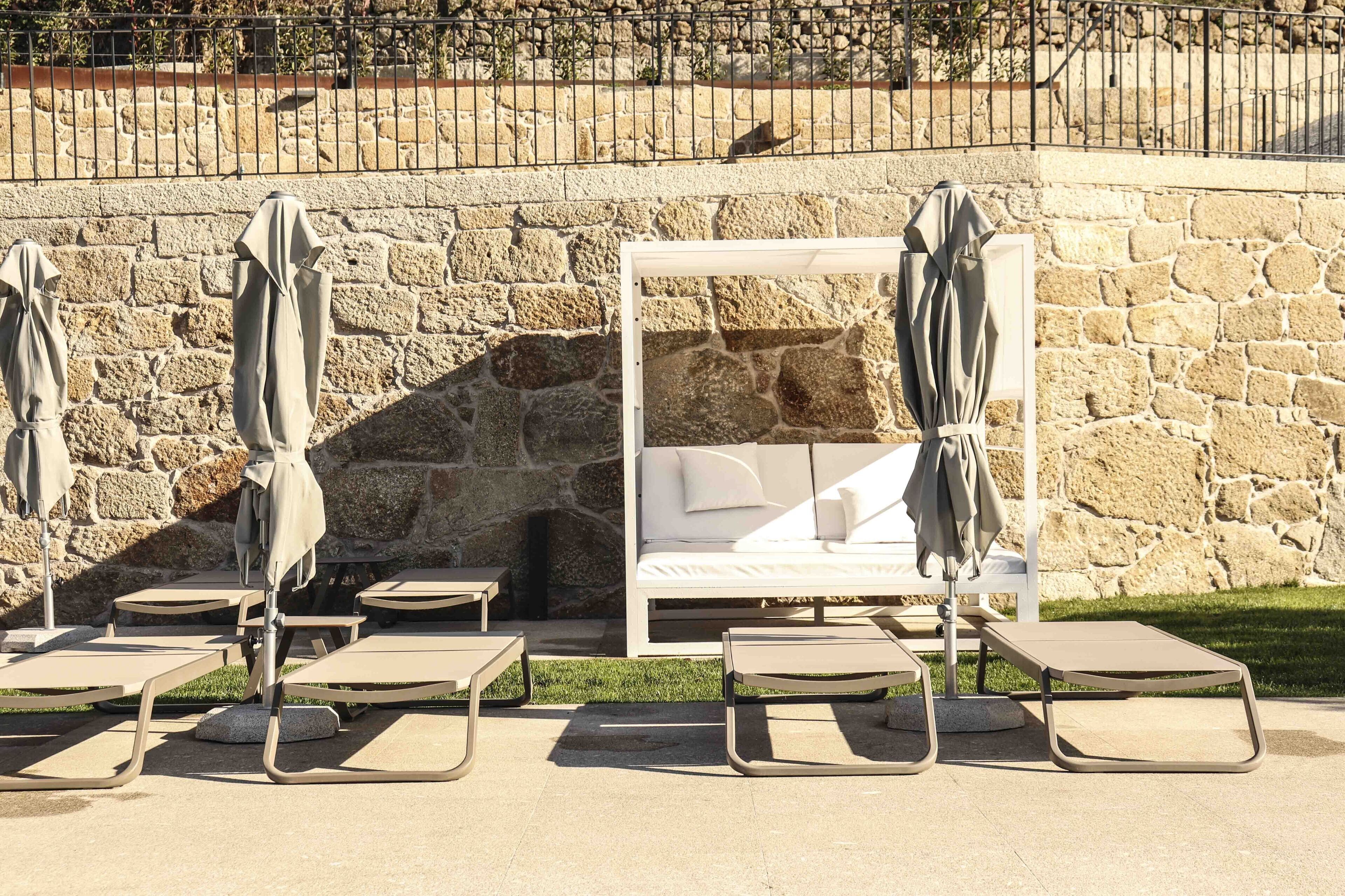 A stone wall with a white bed and lounge chairs with umbrellas.