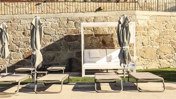 Un muro de piedra con una cama blanca y sillones de descanso con sombrillas.