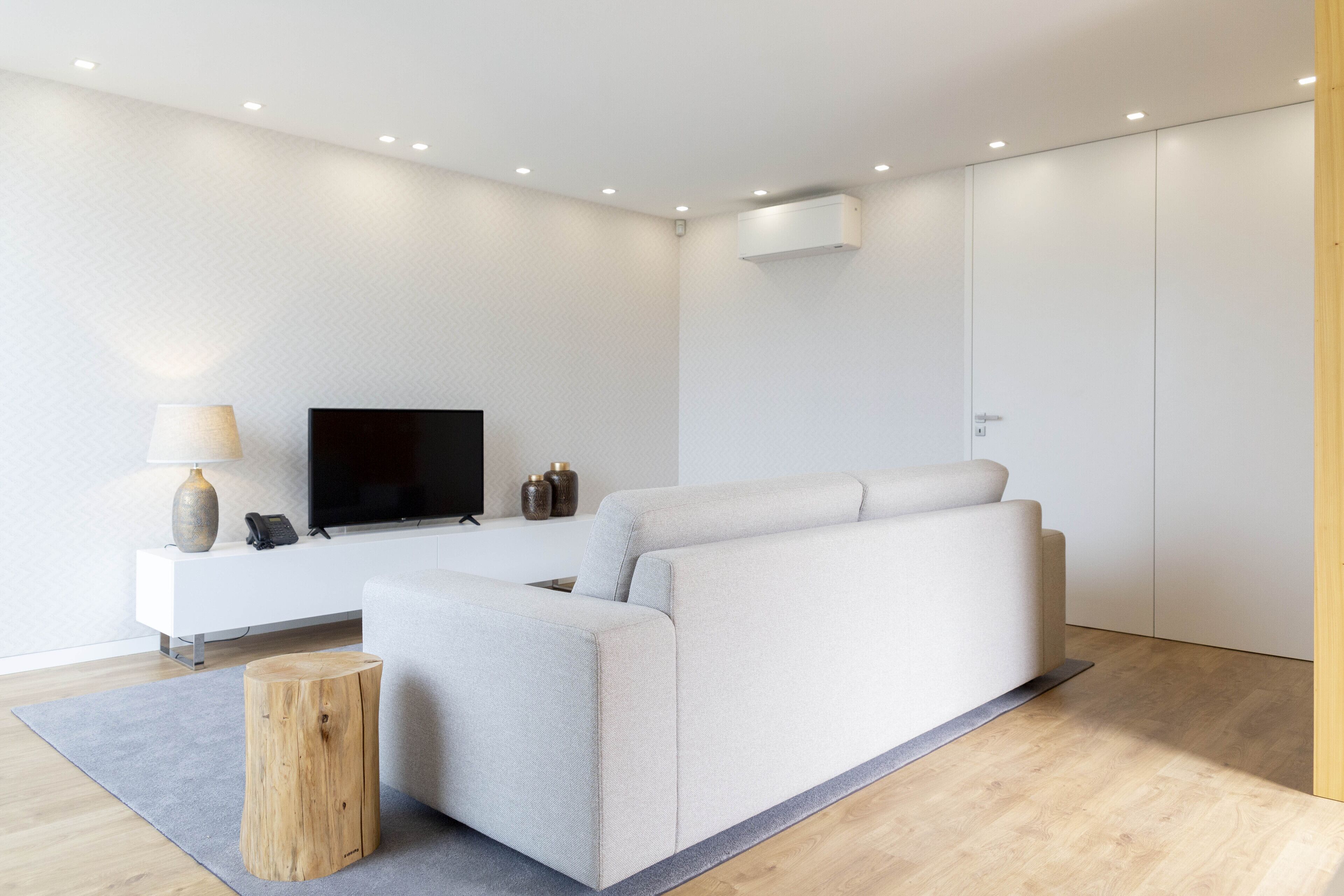 A modern living room with a sofa, a flat-screen TV, a small side table, and a wooden stool.