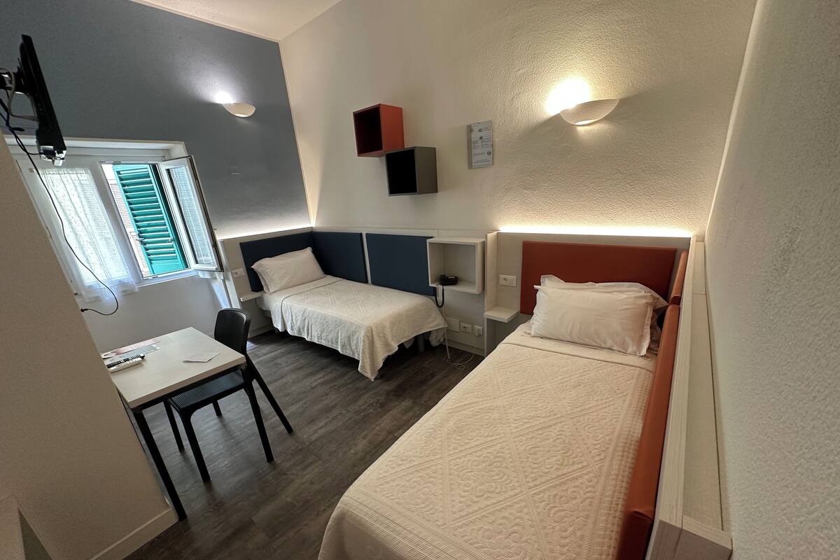 A small, well-lit room with two single beds, a desk, and a chair. There is a window with blinds, a wall-mounted shelf with a phone, and a wall-mounted light fixture.