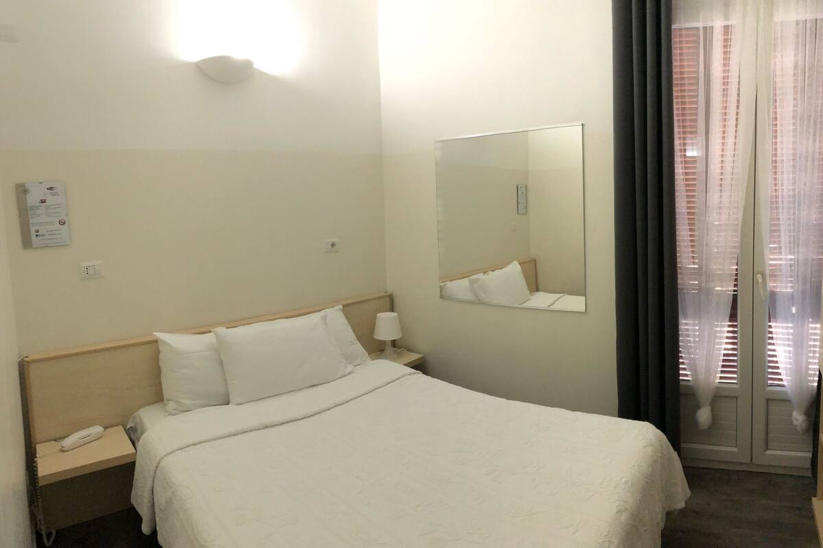 A hotel room with a bed, a bedside table with a lamp, a mirror, and a window with blinds.