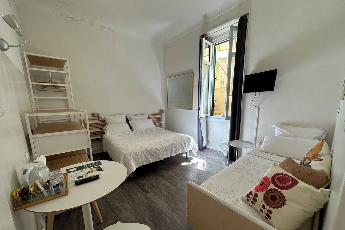 A compact hotel room with two single beds, a small round table, a chair, a desk with a computer and a lamp, and a window with curtains.