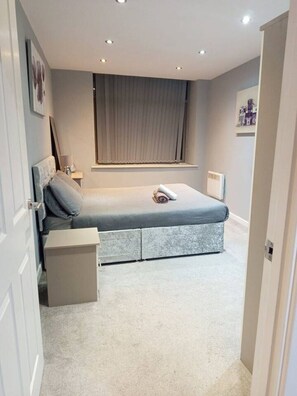Un dormitorio con una cama, una mesita de noche, cortinas en la ventana y una puerta.