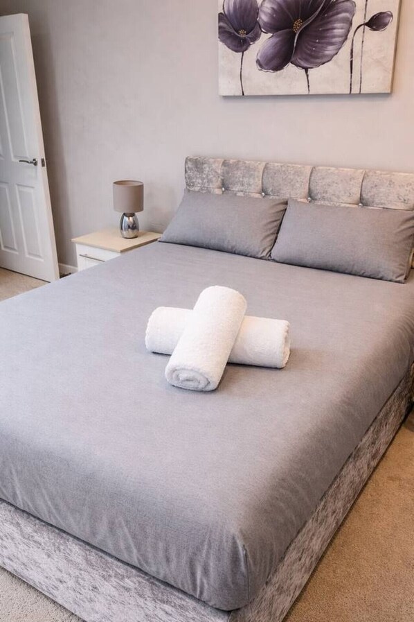 Una cama bien hecha con ropa de cama gris y toallas blancas enrolladas, una mesita de noche con una lámpara, y una pared con un cuadro floral.