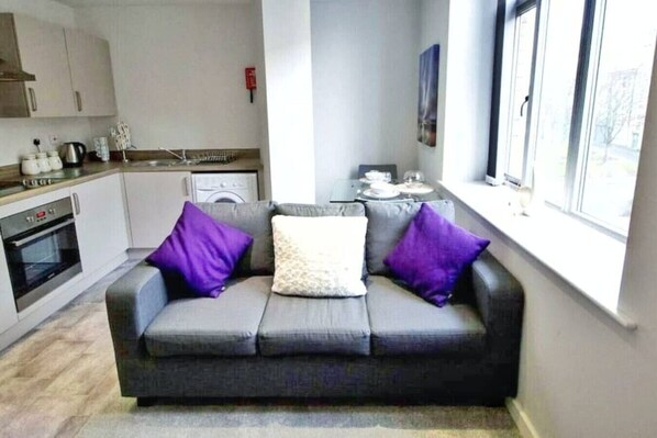 A modern kitchen with a built-in oven, a dining table with chairs, and a gray sofa with purple cushions.