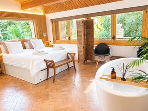 Ein Schlafzimmer mit einem Bett, einer Bank, einem Sessel, einer Badewanne und einem Fenster mit Blick auf Grünflächen.