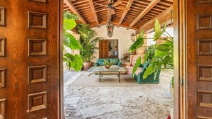 Une porte en bois ouverte donne sur un vaste patio doté d’un sol en pierre, d’un ventilateur de plafond et de divers pots de plantes.