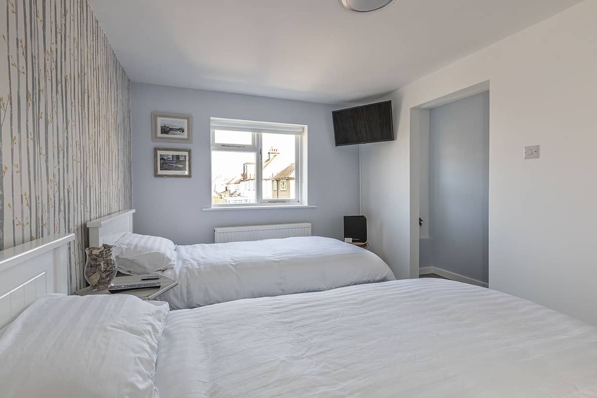 A bedroom with two beds, a window with a view of buildings, a television on the wall, and a radiator.