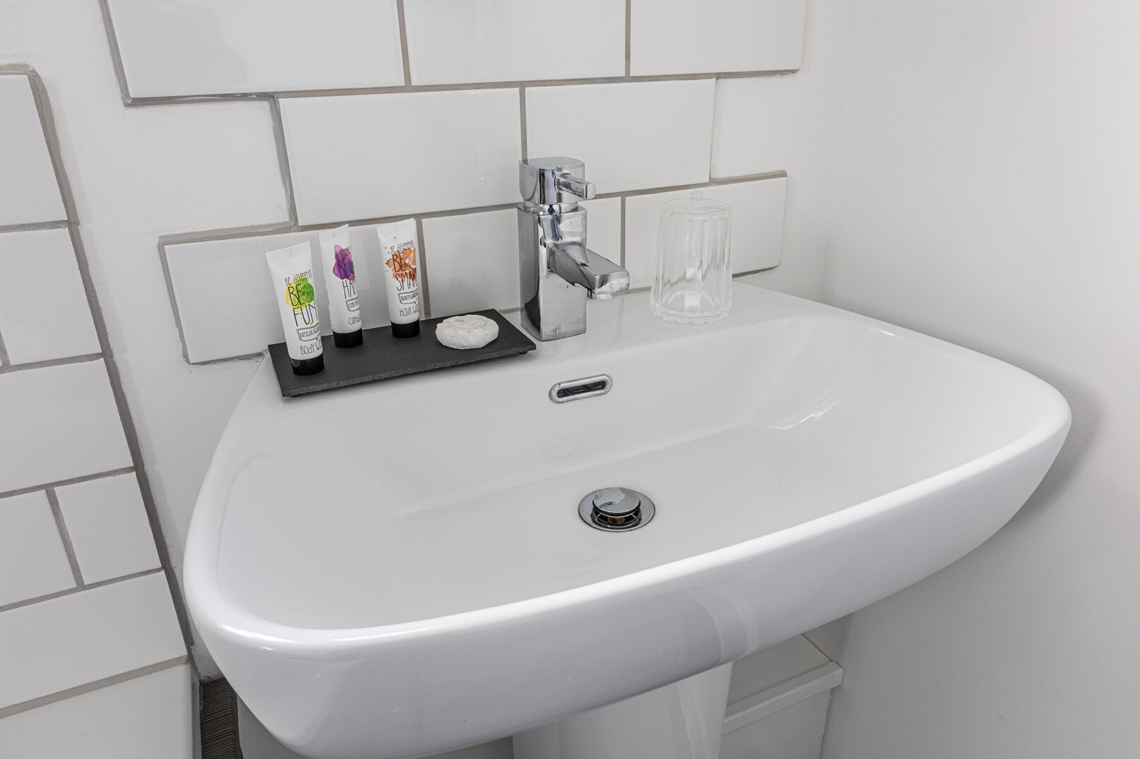 A white bathroom sink with a chrome faucet, a glass, and three bottles of soap on a black tray.