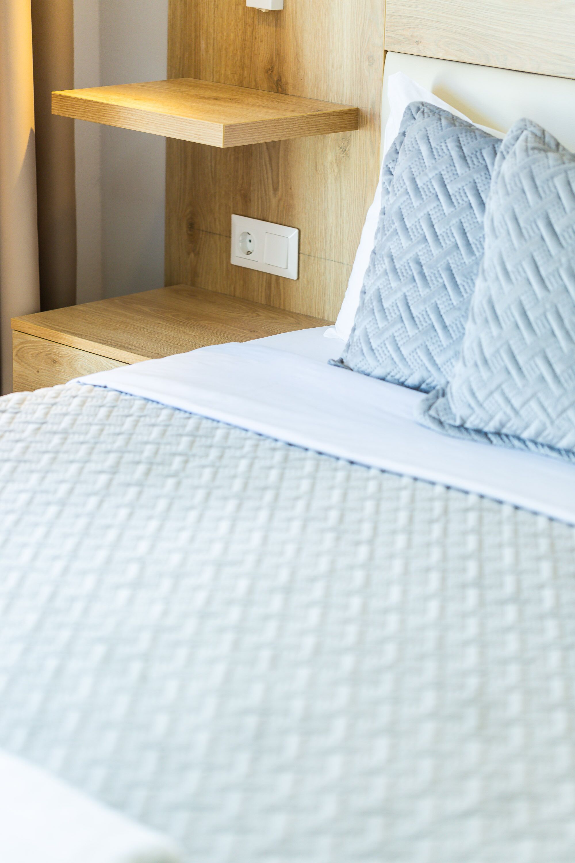Una cama con cubrecolcha blanco acolchado, dos almohadas azul claro y un estante de madera encima.