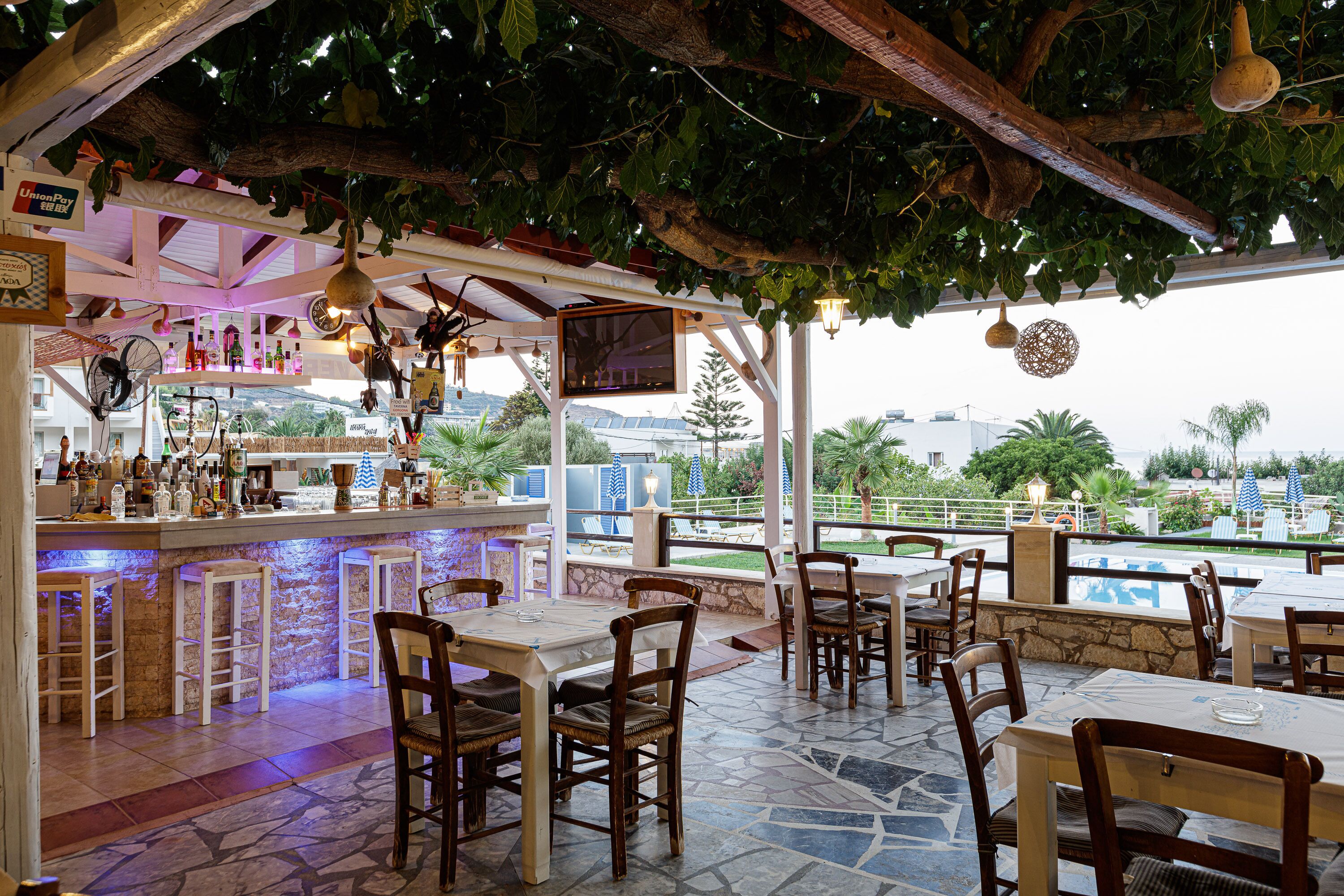 Een openluchtrestaurant met stenen vloeren, houten tafels en stoelen, een bar met flessen en uitzicht op een zwembad en groen.