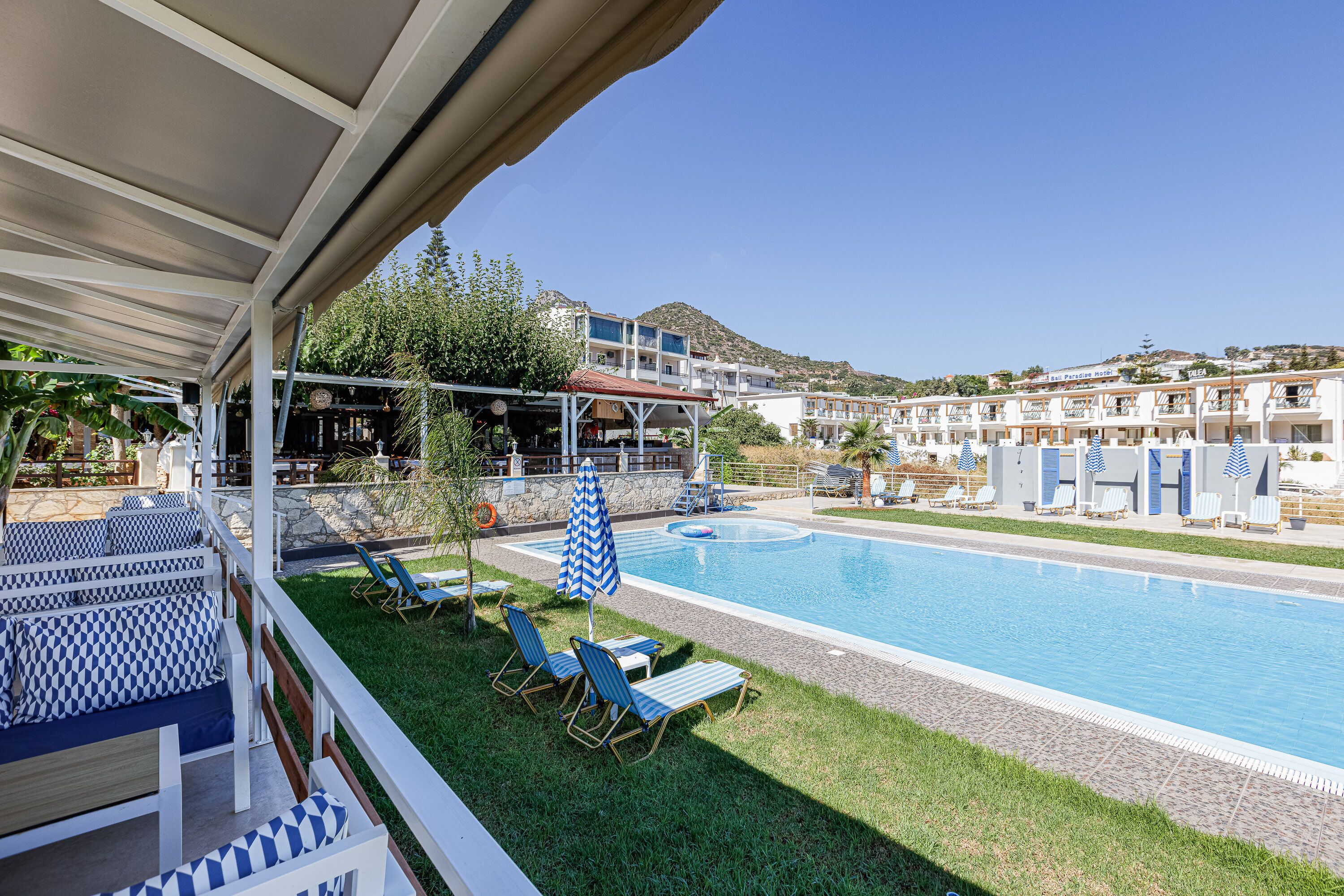 A pool area with lounge chairs and umbrellas, a covered seating area, and a view of hotel buildings in the background.