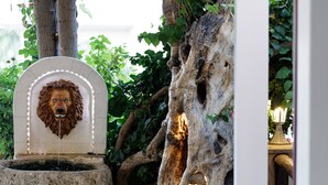 A stone fountain with a lion head sculpture, surrounded by greenery and a large tree trunk.