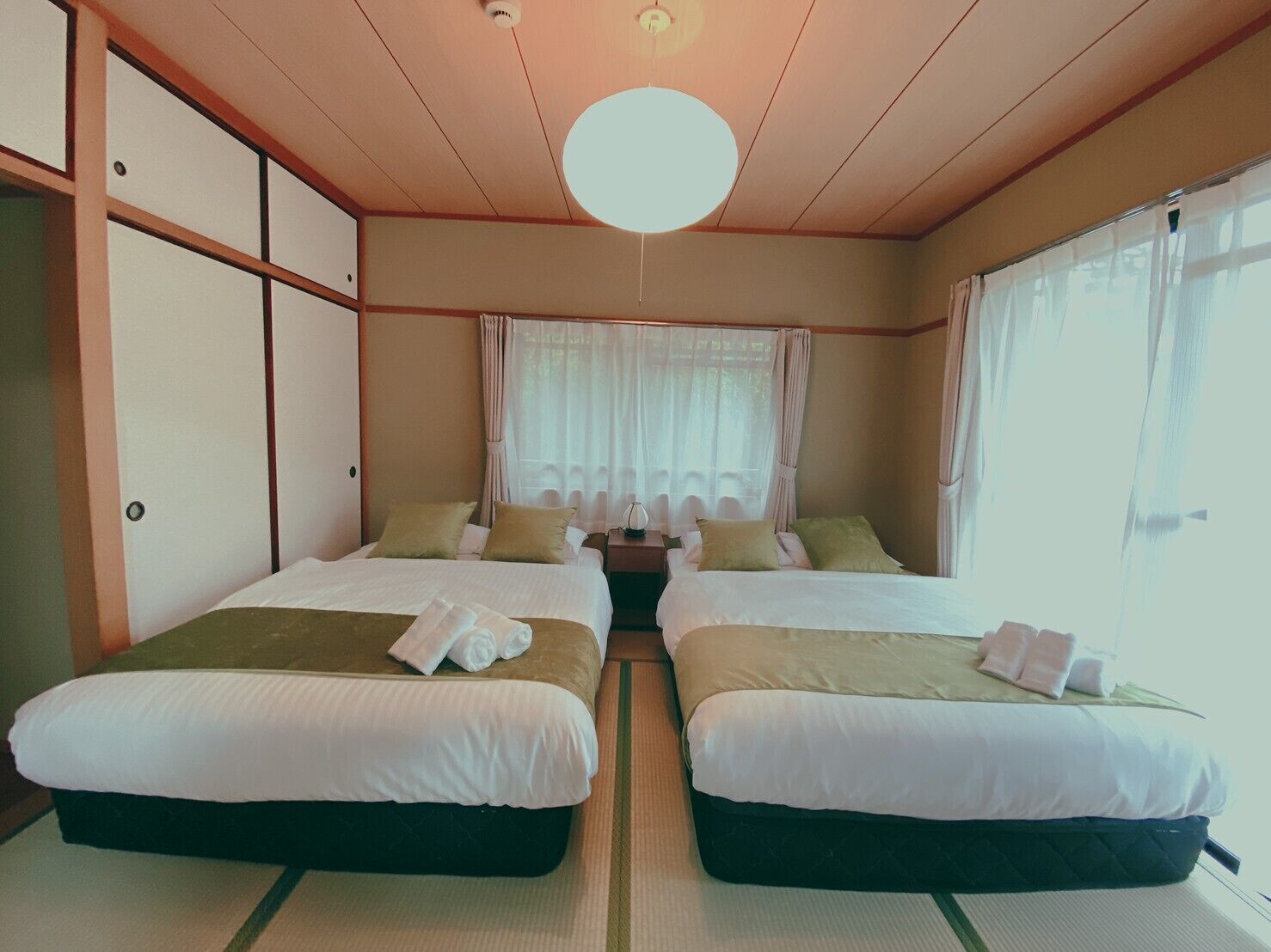 Habitación de hotel con dos camas, una lámpara en el techo, una ventana con cortinas y una mesita de noche.