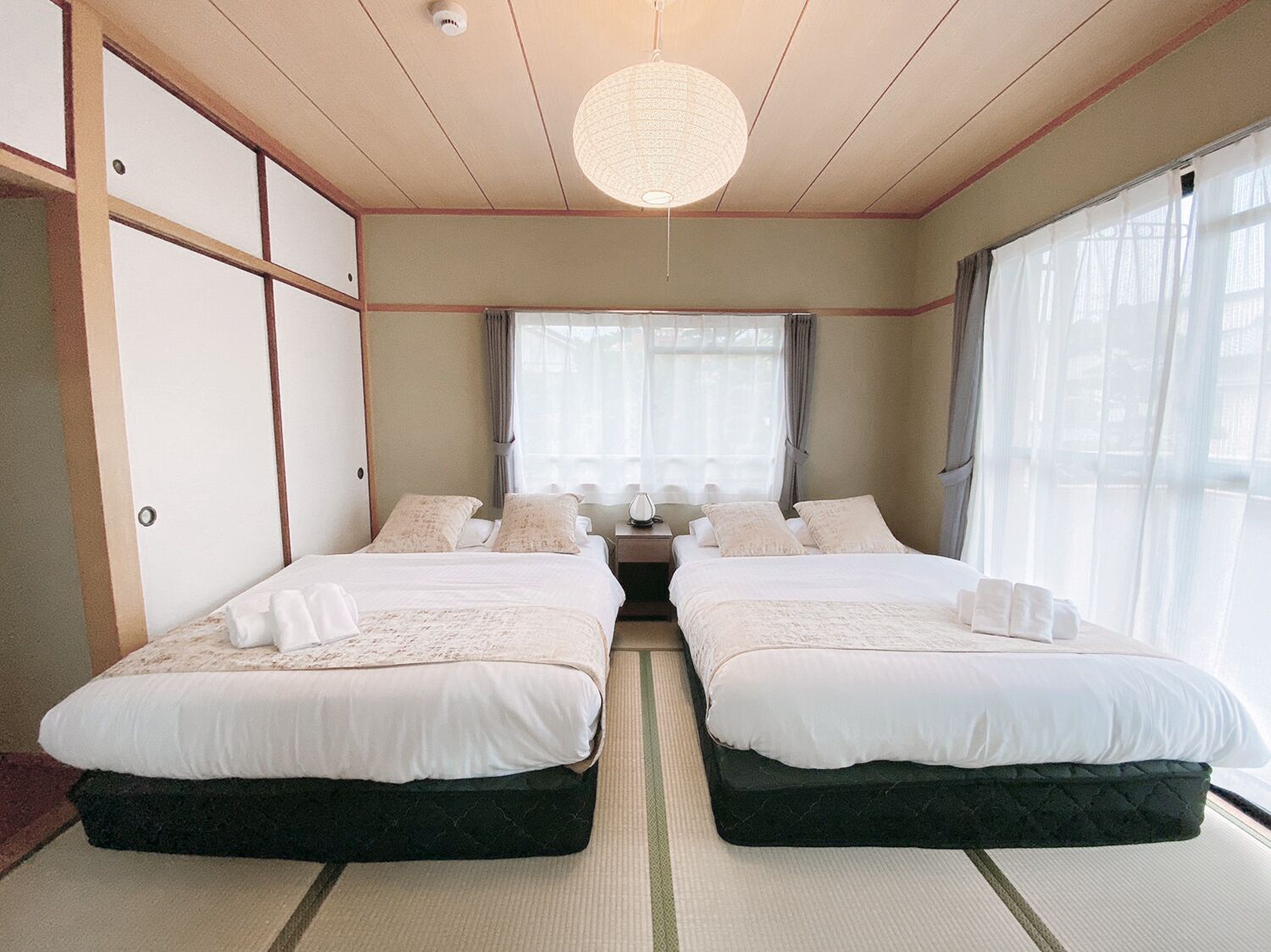 A traditional Japanese-style bedroom with two beds, a tatami mat floor, a hanging light fixture, and a view of the outside through large windows.