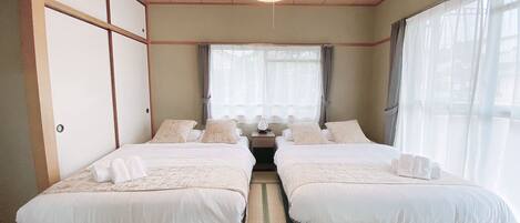 A traditional Japanese-style bedroom with two beds, a tatami mat floor, a hanging light fixture, and a view of the outside through large windows.
