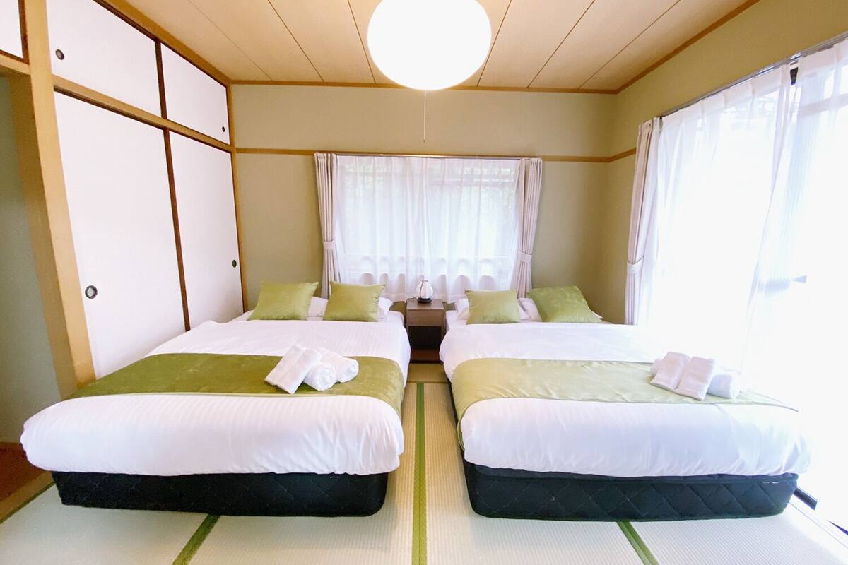 A hotel room with two beds, a bedside table, a lamp, and a window with sheer curtains.