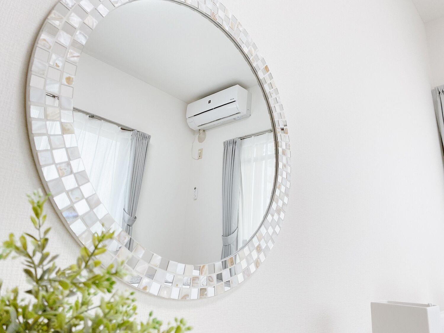 A round mirror with a mosaic border, a ceiling-mounted air conditioner, and a plant in the foreground.