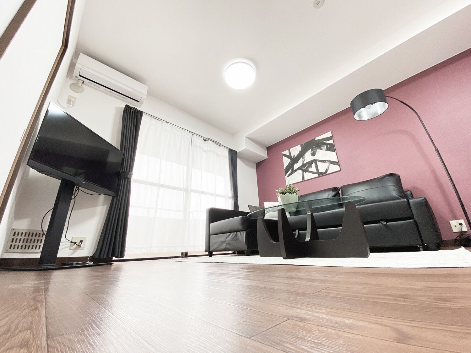 A modern living room with a black sofa, a glass coffee table, and a large window with sheer curtains.