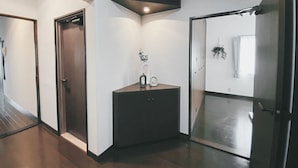 A hallway with a dark wooden floor, white walls, and a glass door leading to a room. There is a small table with a plant and a white vase. - OUCHI HOTEL Itsukaichi (Hiroshima)