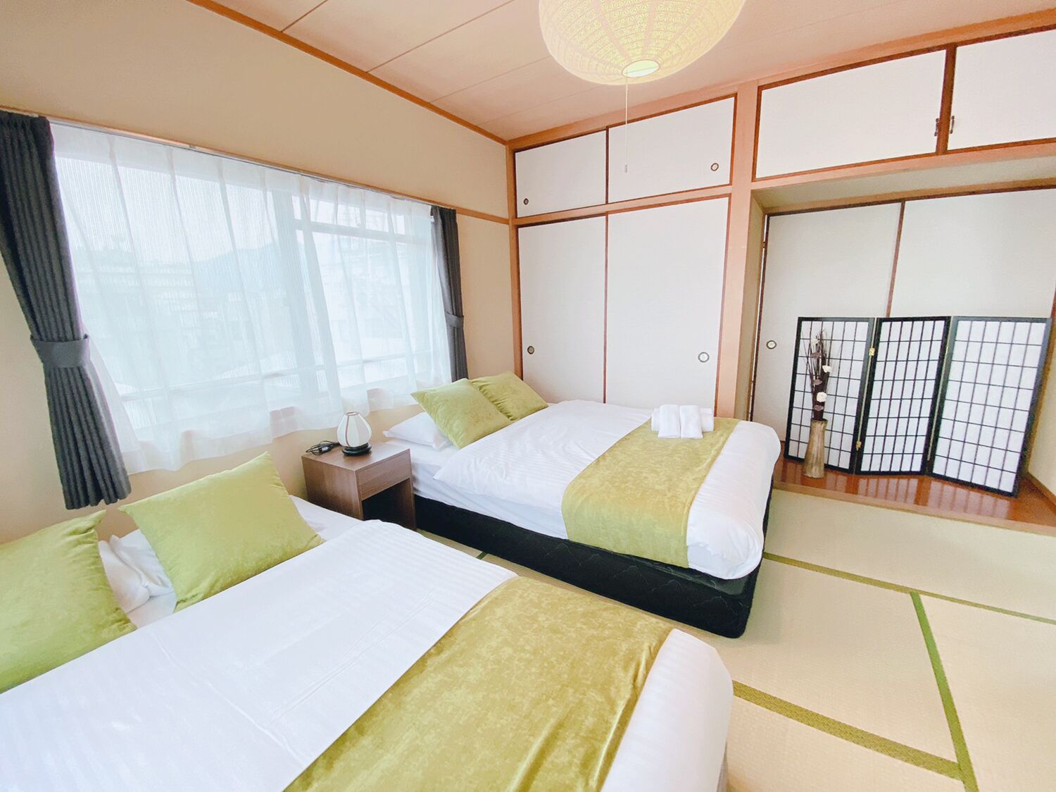 A hotel room with two beds, a bedside table, a lamp, a window with curtains, and a traditional Japanese sliding door.