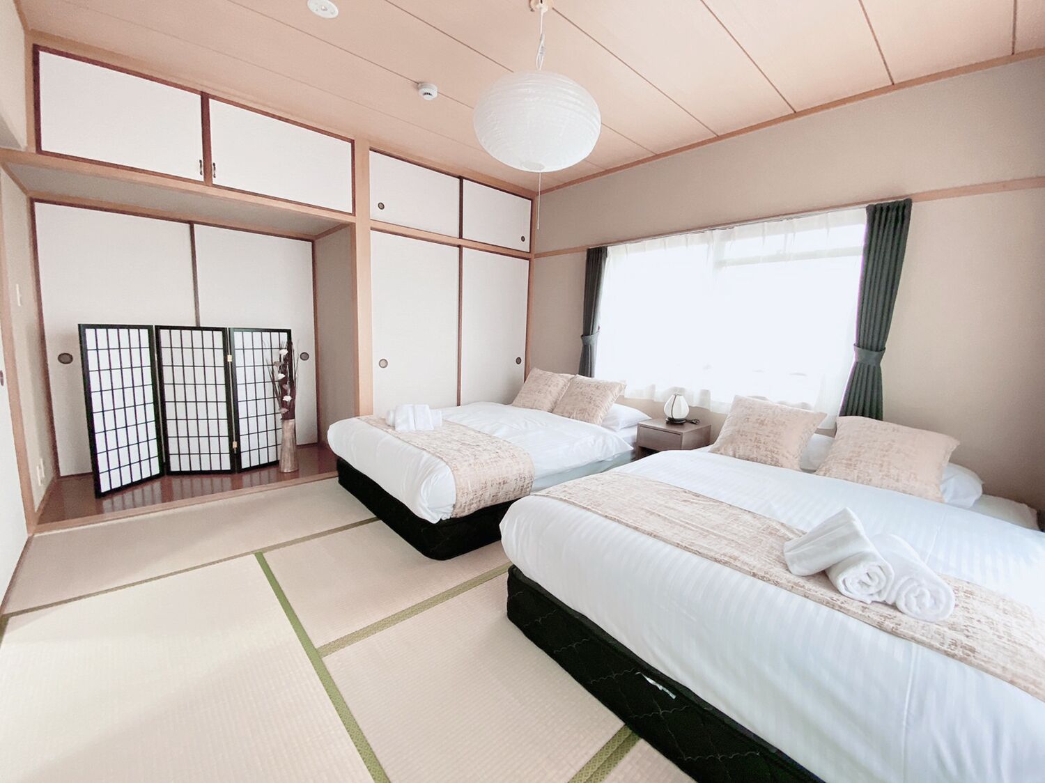 A traditional Japanese-style bedroom with tatami flooring, two beds with white bedding, a shoji screen, and a hanging paper lantern.