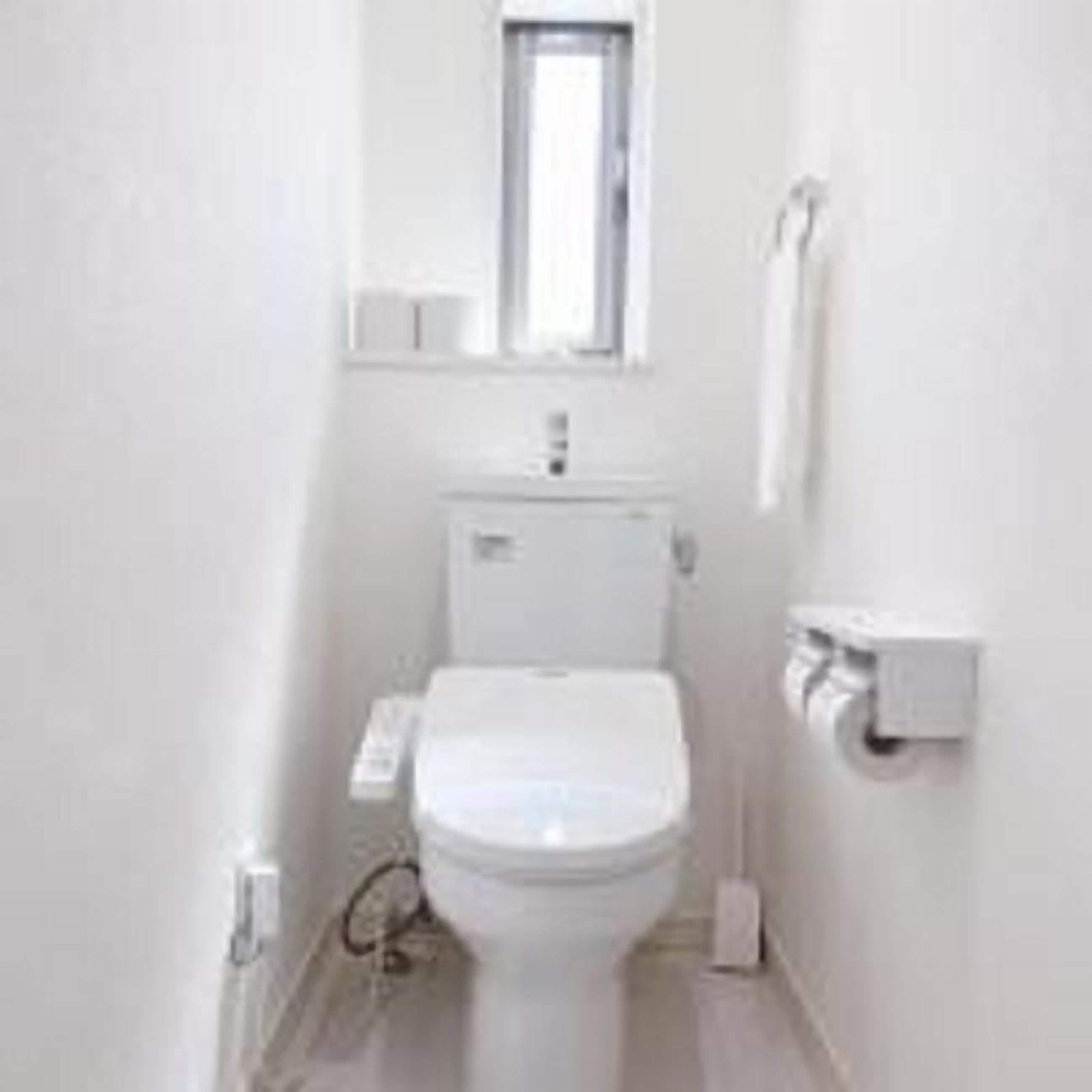 A small, white bathroom with a toilet, a window above the sink, and a towel dispenser on the wall.