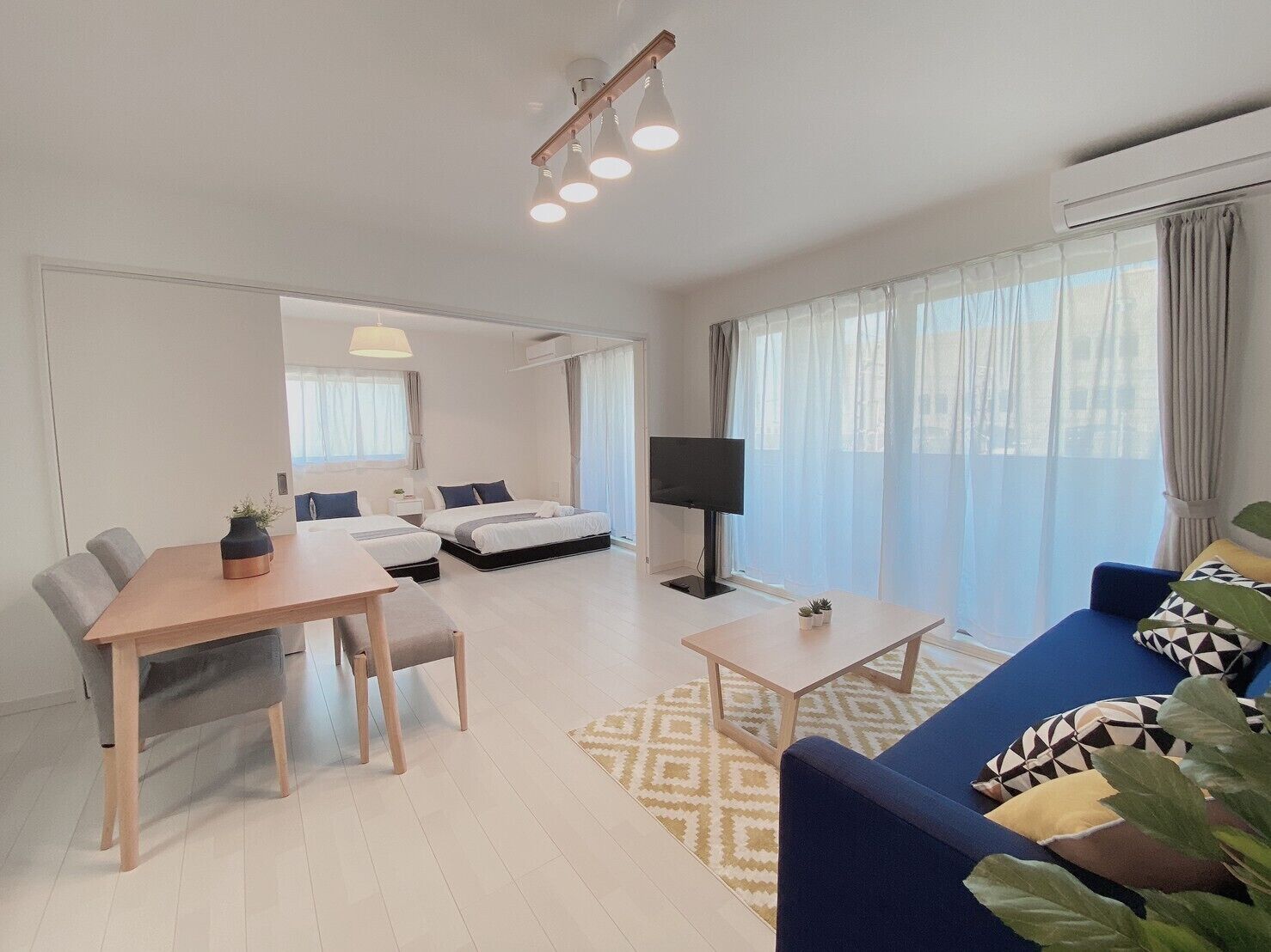 Una sala de estar moderna y bien iluminada con comedor, televisor y un amplio ventanal con cortinas.