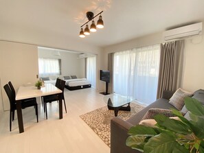 A modern, well-lit hotel room with a dining area, a sofa, a TV, and a large window with curtains. - OUCHI HOTEL Miyajimaguchi (Hatsukaichi)