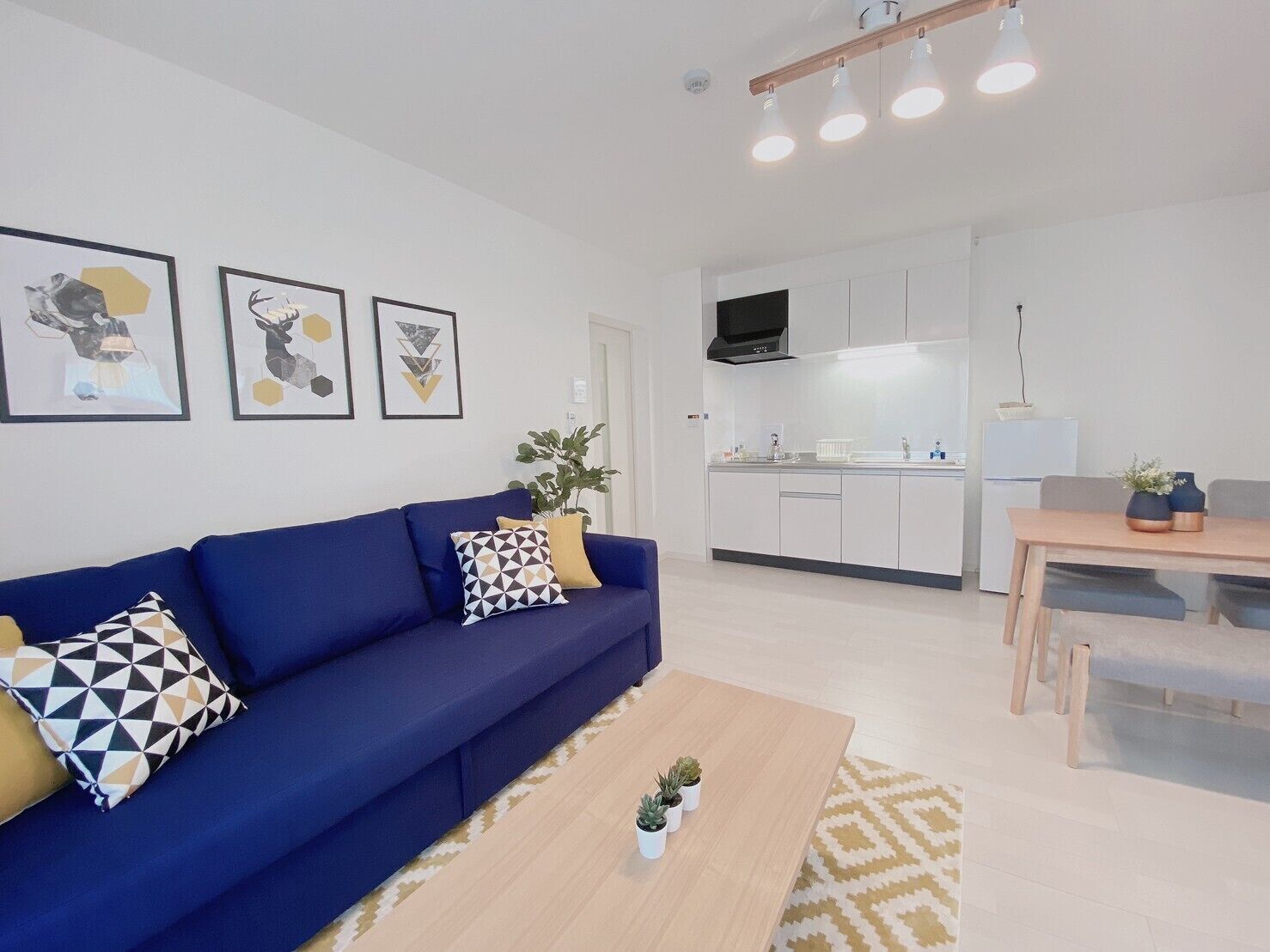 Una sala de estar moderna con un sofá azul, una mesa de centro y una zona de comedor con una mesa y sillas de madera. Se observa una cocina al fondo con armarios blancos y una campana extractora negra.