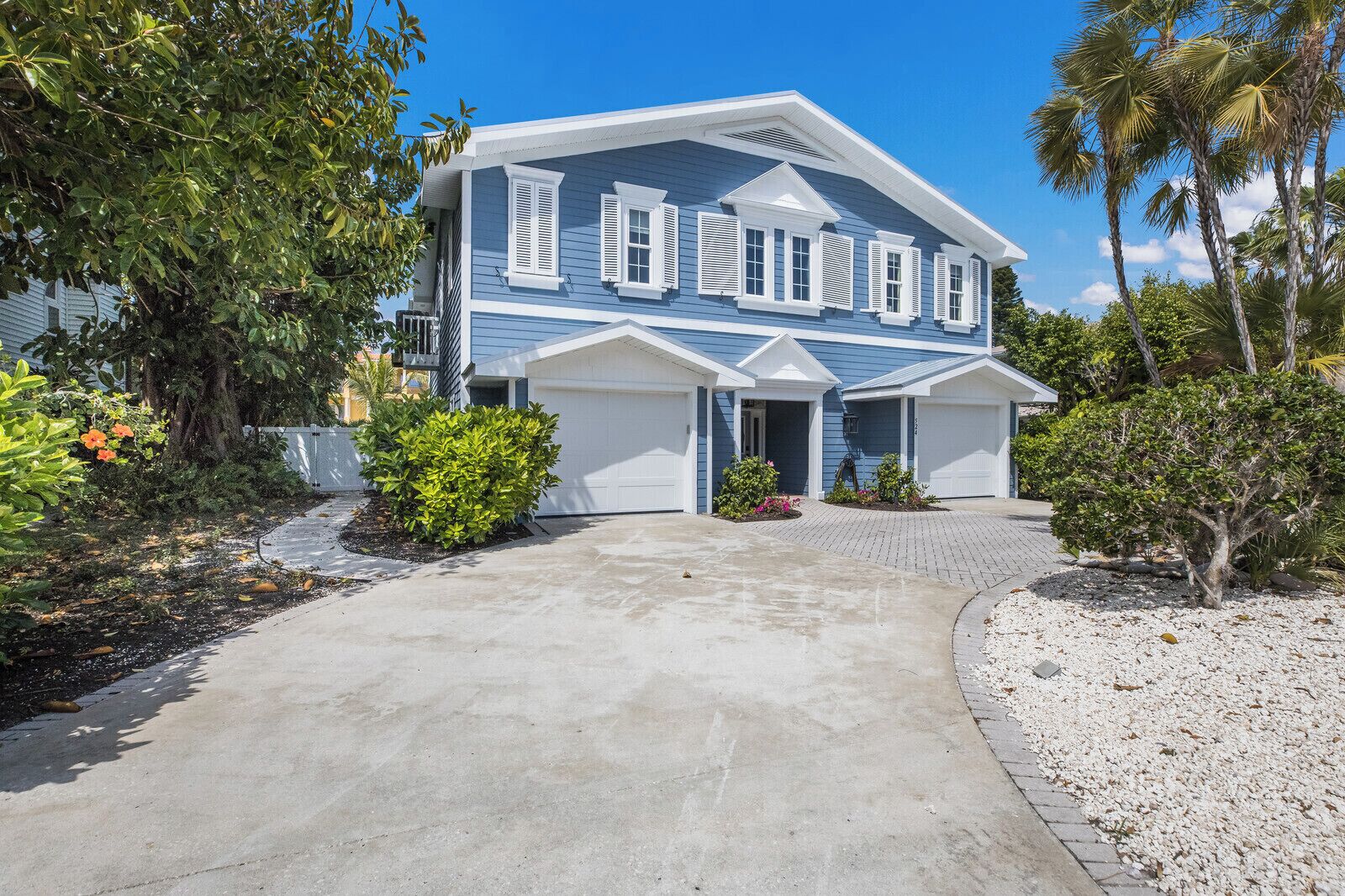 A two-story house with a front garage, surrounded by tropical vegetation and a paved driveway.