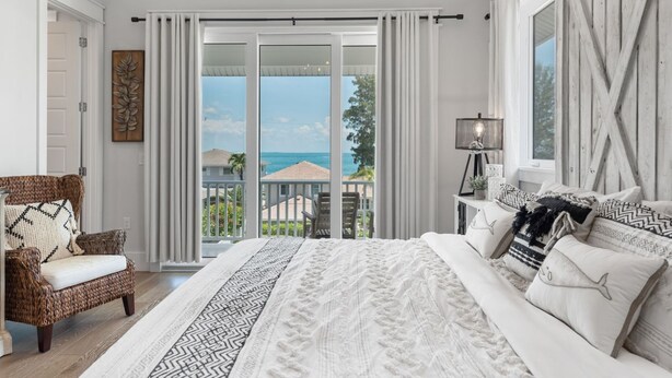 A bedroom with a large bed, a chair, a nightstand, and a view of the ocean through a sliding glass door. - Funky Beach Farmhouse: Beautiful Bay views, heated pool, hot tub, lounge area (Anna Maria)