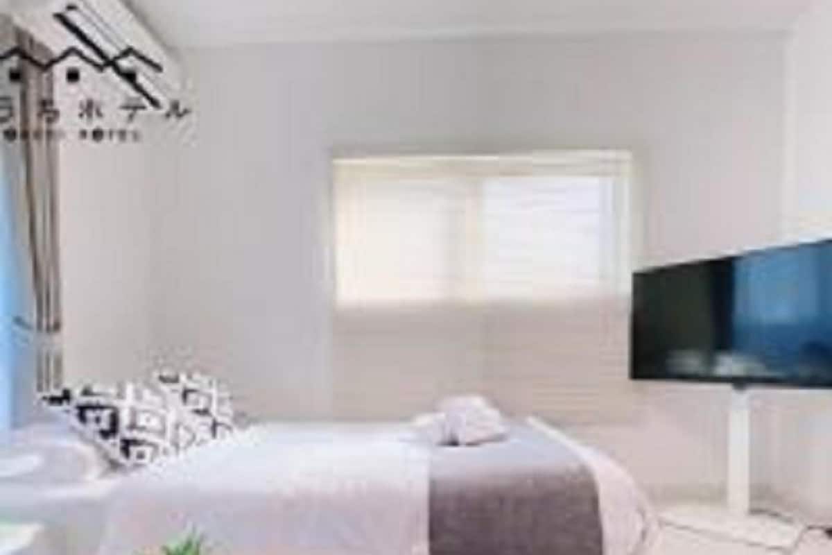 Ein modernes Hotelzimmer mit Bett, Fernseher, kleinem Tisch mit Pflanze und Fenster mit Jalousien.
