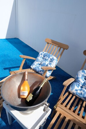 Una silla de madera con un cojín floral, un cuenco metálico con una botella de champán y una mesa blanca.